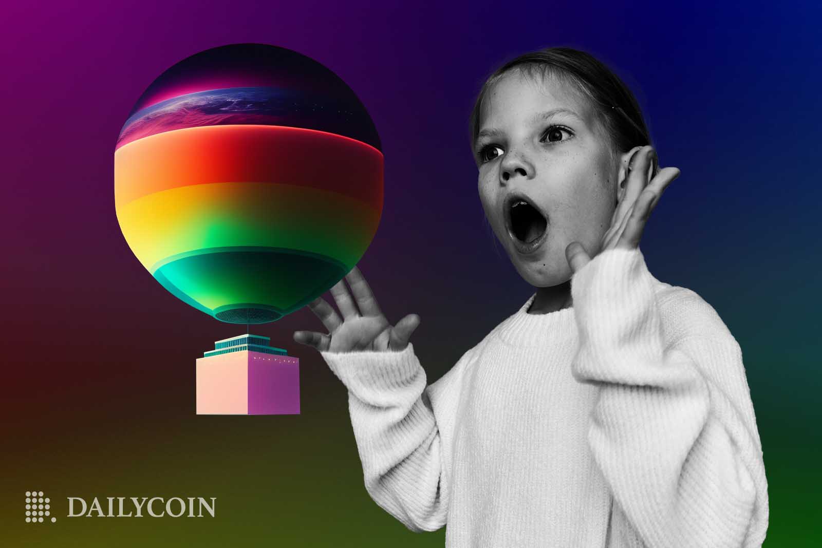 A girl with her mouth open and hands up looks at a colorful air balloon