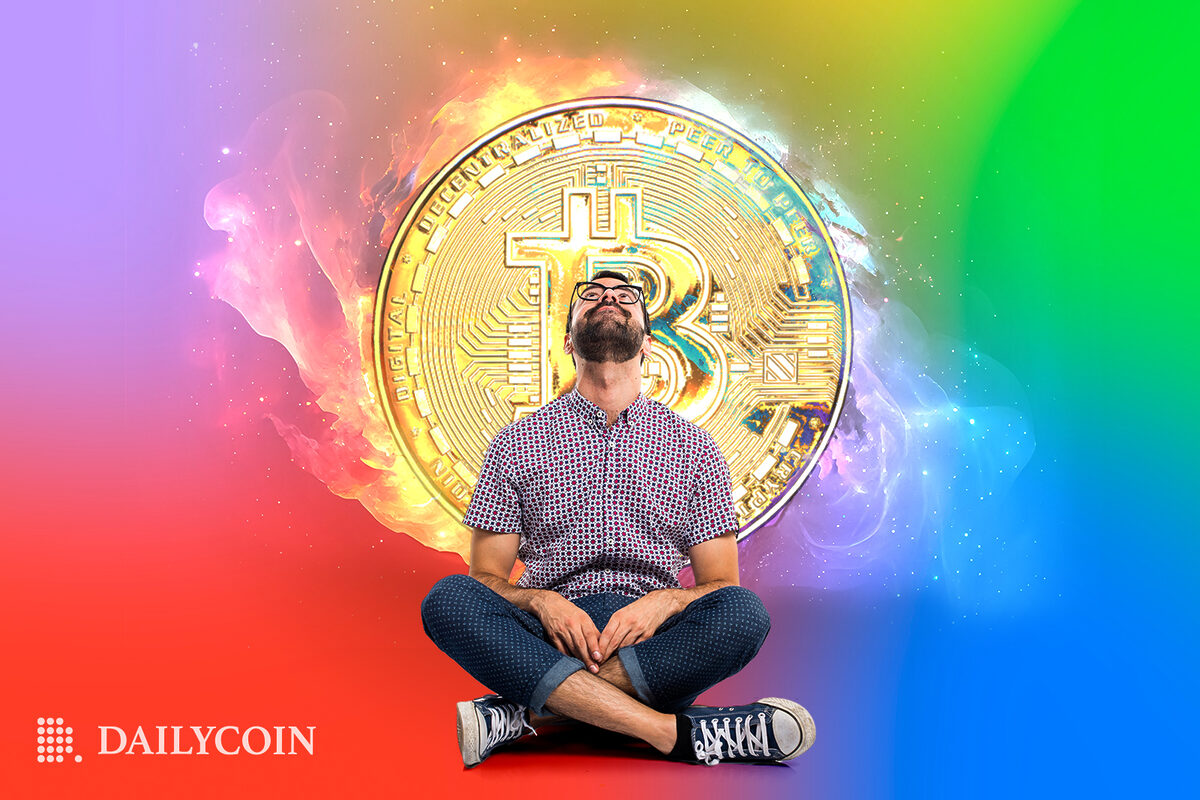 Man sitting cross legged looking up, with a giant bitcoin behind him surrounded by a rainbow coloured background.