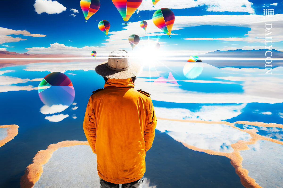 Man in a yellow jacket stands in front of a lake with colorful air balloons landing in the background.