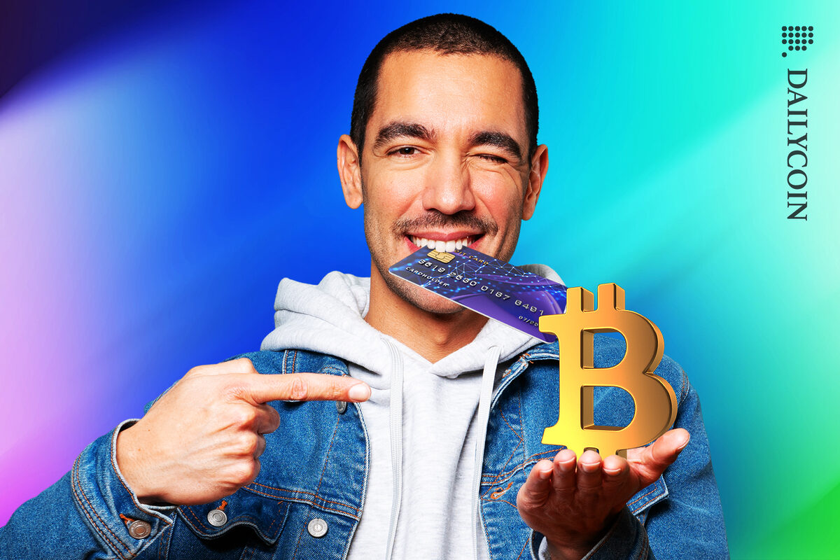 Man bitting on his credit card smiling. His one hand finger is pointing to the other holding a Bitcoin logo sculpture.