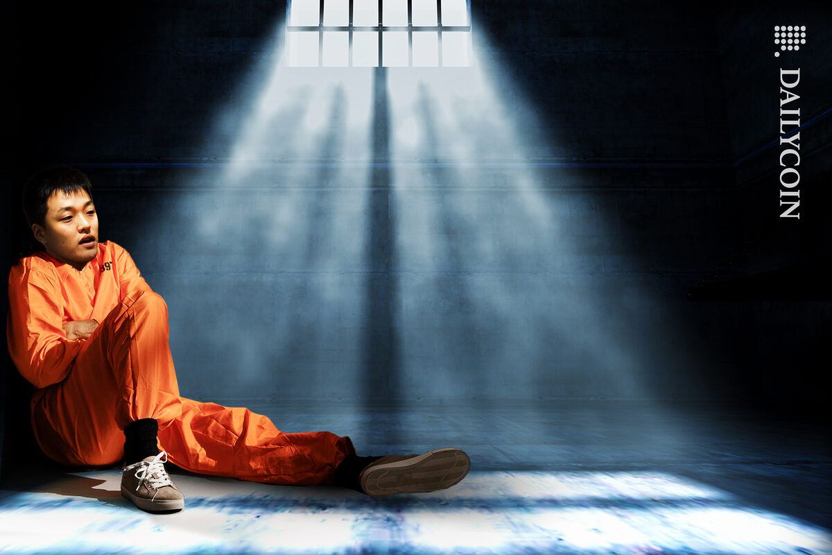 Do Kwon with arms crossed is wearing an orange prison suit and is sitting on the floor in the corner of a prison cell.