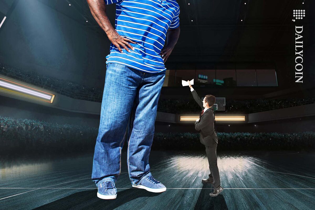 Tiny man in suit trying to hand some documents to Shaq, but he cant reach up high enough.
